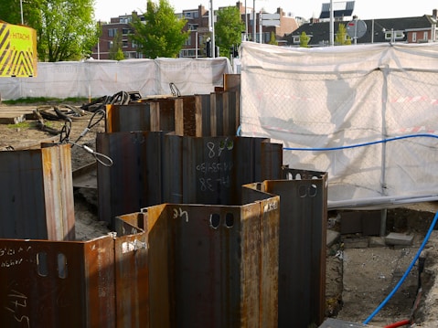 A construction site with metal sheet piles arranged in a circle in the foreground. The background shows a fenced-off area with a white tarp and a 'Hitachi' branded sign. Buildings and trees are visible in the distance.