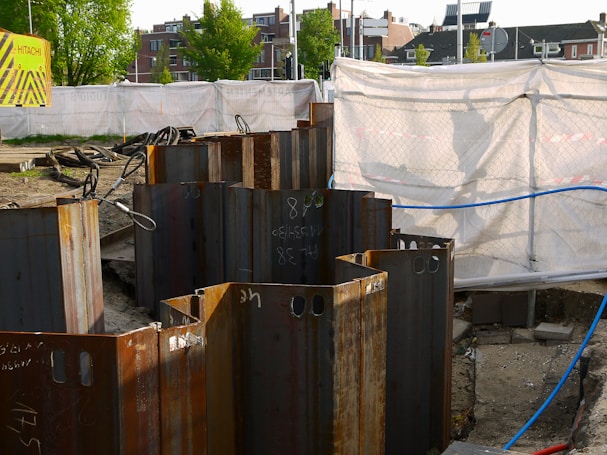 A construction site with metal sheet piles arranged in a circle in the foreground. The background shows a fenced-off area with a white tarp and a 'Hitachi' branded sign. Buildings and trees are visible in the distance.