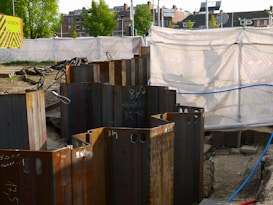A construction site with metal sheet piles arranged in a circle in the foreground. The background shows a fenced-off area with a white tarp and a 'Hitachi' branded sign. Buildings and trees are visible in the distance.