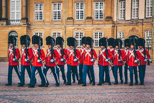 A group of uniformed guards wearing red coats, black trousers, and tall black fur hats marching in formation. The setting appears to be an open courtyard of a historic building with large windows and stonework.