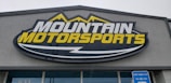 A large, rectangular business sign reads 'Mountain Motorsports' in bold letters. The text 'MOUNTAIN' is white, while 'MOTORSPORTS' is yellow, both set against a dark background. Above the text, there is an outline of mountain peaks in yellow.