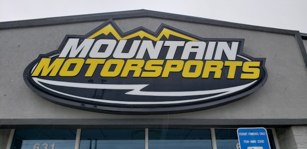 A large, rectangular business sign reads 'Mountain Motorsports' in bold letters. The text 'MOUNTAIN' is white, while 'MOTORSPORTS' is yellow, both set against a dark background. Above the text, there is an outline of mountain peaks in yellow.