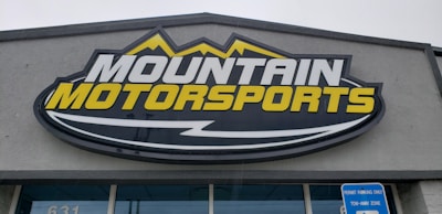 A large, rectangular business sign reads 'Mountain Motorsports' in bold letters. The text 'MOUNTAIN' is white, while 'MOTORSPORTS' is yellow, both set against a dark background. Above the text, there is an outline of mountain peaks in yellow.