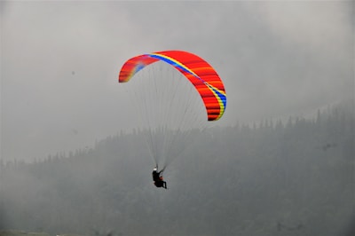 A person is paragliding with a brightly colored parachute against a misty, overcast background. The parachute features vivid stripes of red, yellow, and blue. The backdrop shows a range of dark, hazy mountains with a dense layer of fog, creating a sense of height and adventure.