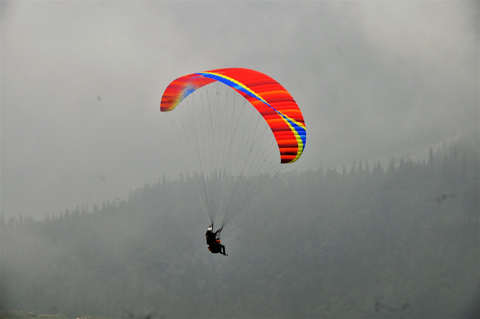 A person is paragliding with a brightly colored parachute against a misty, overcast background. The parachute features vivid stripes of red, yellow, and blue. The backdrop shows a range of dark, hazy mountains with a dense layer of fog, creating a sense of height and adventure.
