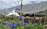 Colorful prayer flags are strung across a natural landscape, with blue wildflowers in the foreground. Several tents are pitched behind a row of wicker chairs and rustic wooden fencing. The scene is set against a backdrop of rugged mountains partially hidden by clouds.