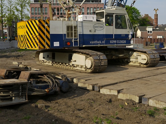 A large construction crane with caterpillar tracks is positioned on a wooden platform, surrounded by construction materials and dirt. The yellow and black striped pattern on the crane's counterweight is prominent. In the background, there are residential buildings and trees under a clear sky.