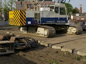A large construction crane with caterpillar tracks is positioned on a wooden platform, surrounded by construction materials and dirt. The yellow and black striped pattern on the crane's counterweight is prominent. In the background, there are residential buildings and trees under a clear sky.