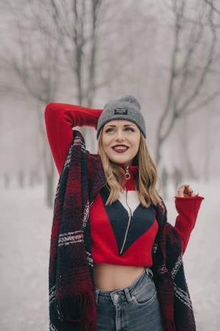 A woman wearing a red and navy sweater with a geometric pattern, a gray beanie, and jeans stands in an outdoor setting surrounded by bare trees. The ground and background are covered with snow, suggesting a winter scene. Her hair is down, and she appears to be smiling with one arm raised behind her head.