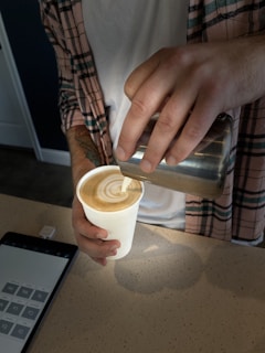 A person wearing a plaid shirt carefully pours steamed milk into a cup of coffee, creating latte art. A tablet with a numeric keypad is placed on the counter nearby.