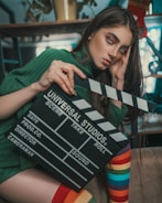 A person with long hair is holding a film clapperboard labeled 'Universal Studios'. They are wearing a green sweater and have rainbow-striped socks. The background features shelves and some decor items.