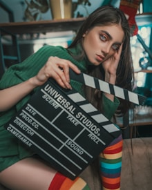 A person with long hair is holding a film clapperboard labeled 'Universal Studios'. They are wearing a green sweater and have rainbow-striped socks. The background features shelves and some decor items.