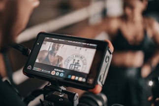 A digital camera screen displays a close-up of a person in athletic wear, possibly recording a fitness video. The person appears to be smiling and is in a well-lit space with a ring-like setting visible in the background.
