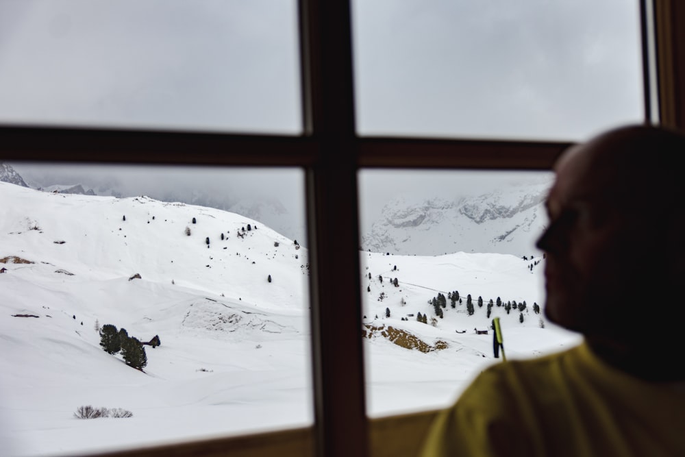 man wearing yellow crew-neck shirt near glass window viewing field covered with snow during daytime