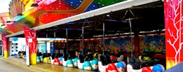 A vibrant and colorful bumper car ride with decorative patterns and lighting, featuring multiple bumper cars lined up and ready for use. The structure above is adorned with colorful artwork, including swirling designs and cosmic imagery.