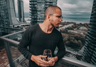 man standing and holding near empty wineglass leaning beside gray framed clear glass building balcony railing at the city during day