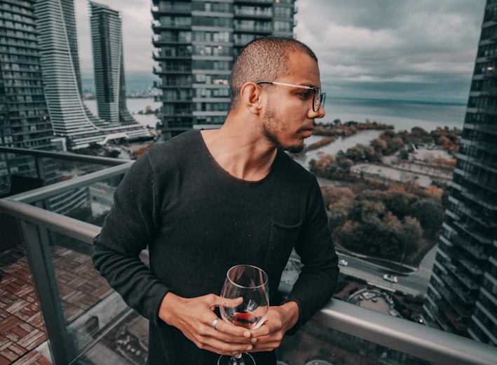 man standing and holding near empty wineglass leaning beside gray framed clear glass building balcony railing at the city during day