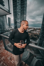man standing and holding near empty wineglass leaning beside gray framed clear glass building balcony railing at the city during day