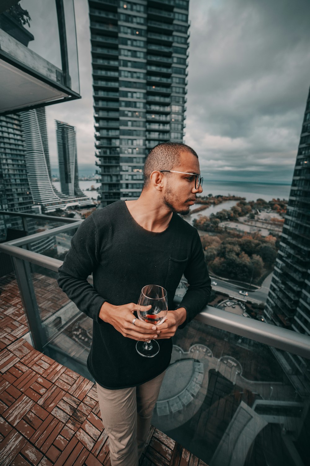 man standing and holding near empty wineglass leaning beside gray framed clear glass building balcony railing at the city during day