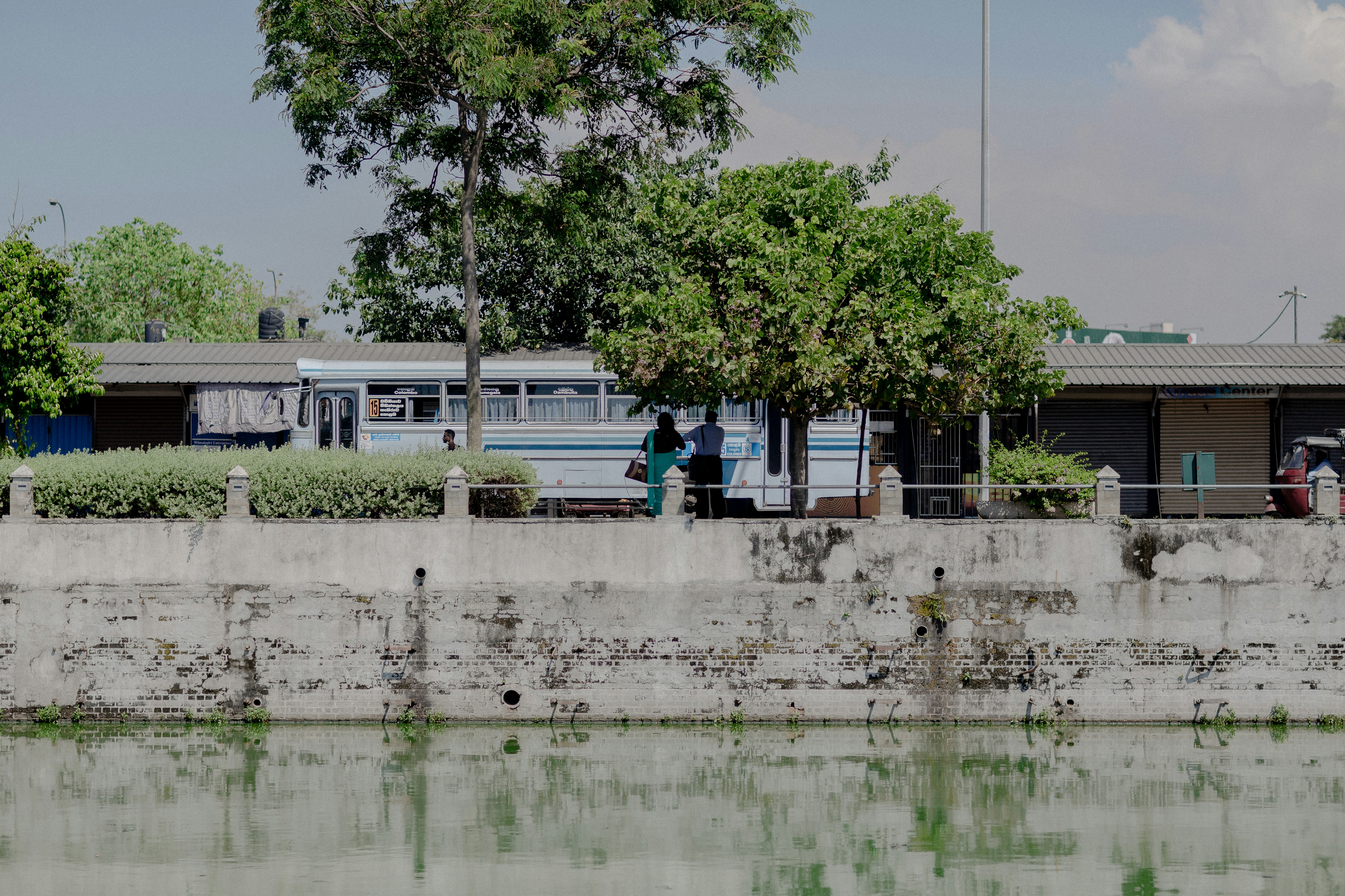 two people standing near white and blue bus beside buildings surrounded with green trees under white and blue sky - Colombo