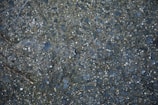 A close-up view of a textured asphalt surface with scattered small stones and pebbles embedded in it. A dry twig can be seen lying on the surface, and the colors range from dark gray to bluish tones.