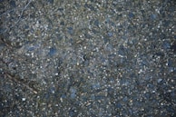 A close-up view of a textured asphalt surface with scattered small stones and pebbles embedded in it. A dry twig can be seen lying on the surface, and the colors range from dark gray to bluish tones.