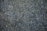 A close-up view of a textured asphalt surface with scattered small stones and pebbles embedded in it. A dry twig can be seen lying on the surface, and the colors range from dark gray to bluish tones.