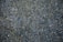A close-up view of a textured asphalt surface with scattered small stones and pebbles embedded in it. A dry twig can be seen lying on the surface, and the colors range from dark gray to bluish tones.