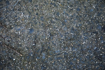 A close-up view of a textured asphalt surface with scattered small stones and pebbles embedded in it. A dry twig can be seen lying on the surface, and the colors range from dark gray to bluish tones.