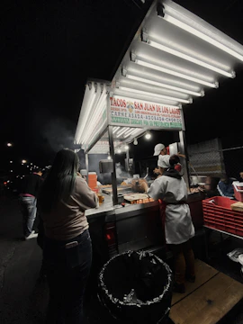 Close-up shot of sizzling tacos being served at a vibrant Long Beach street food stall at sunset.
