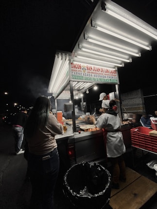 A night-time street food scene with a taco stand illuminated by bright fluorescent lights. A cook wearing a white apron and cap is preparing food on a grill, while a customer stands by the counter. The stall has a sign that reads 'Tacos San Juan de Los Lagos' offering various meat options.
