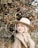 smiling woman wearing brown coat and beige hat standing near flowering tree