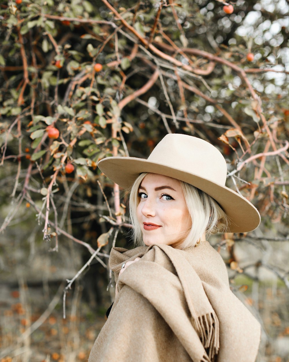 smiling woman wearing brown coat and beige hat standing near flowering tree