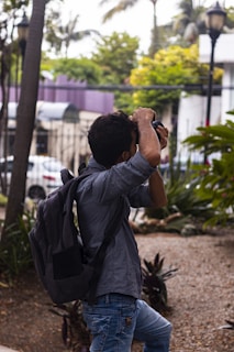A person with dark hair is holding a camera up to their face, appearing to take a photograph. They are wearing a gray checkered shirt and faded blue jeans, with a black backpack on their back. The setting appears to be an outdoor garden area with young trees and various plants around, and a purple building is visible in the background.