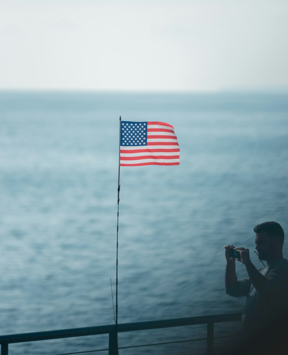 man standing while taking photo near railings viewing body of water and waving United States of America flag