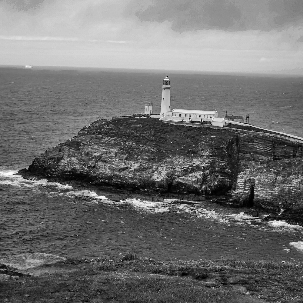 grayscale photography of lighthouse on cliff viewing body of water