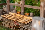A traditional Japanese water basin setup, featuring bamboo rods and a wooden ladle placed on top. The structure is surrounded by a wooden fence and lush green plants. The netted base adds texture to the earthy and natural ambiance.