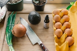 A wooden cutting board holds fresh ingredients including a bunch of green onions, a round yellow onion, and a dark avocado. A chef's knife with a wooden handle lies beside them. Nearby, a carton contains several brown eggs, and a small dropper bottle is positioned next to the avocado. In the background, potted plants add a touch of green.