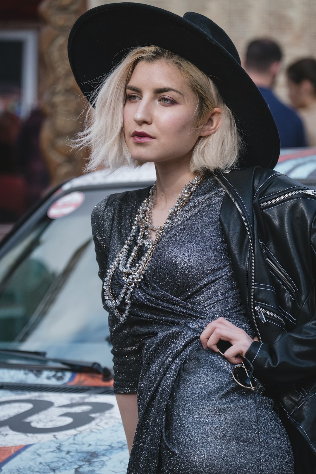 woman wearing black leather zip-up jacket, black hat, and gray long-sleeved dress standing with left hand akimbo near gray vehicle