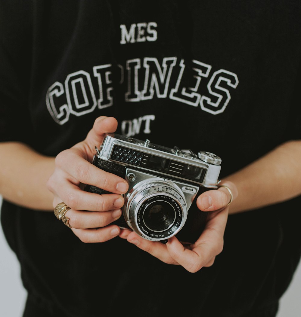 person wearing black and white printed shirt standing while holding black and silver bridge camera