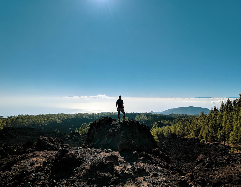 Mann, der auf einem Felsen steht und einen Berg unter blauem und weißem Himmel betrachtet