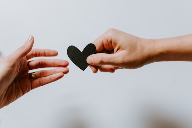 Two hands reaching towards each other, one holding a small black heart-shaped object, symbolizing connection or offering.