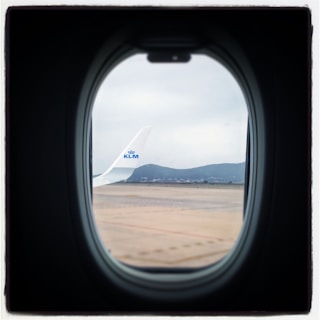 View from an airplane window showing the wingtip with the KLM logo. The scene includes a cloudy sky and distant mountains beyond the airport tarmac.