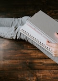 A person sitting with crossed legs wearing light gray jeans holds a book with a gray cover and a spiral notebook. The book has the title 'Love & Respect' on its spine. The surface underneath is made of dark, rustic wood.