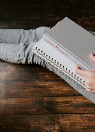 A person sitting with crossed legs wearing light gray jeans holds a book with a gray cover and a spiral notebook. The book has the title 'Love & Respect' on its spine. The surface underneath is made of dark, rustic wood.