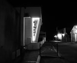 A neon-lit cinema sign is mounted on the side of a building at night. The street is dimly lit with a few street lamps in the background, creating a quiet and deserted atmosphere. The buildings along the street are mostly dark, with just a few lights illuminating them.