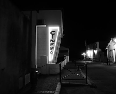 A neon-lit cinema sign is mounted on the side of a building at night. The street is dimly lit with a few street lamps in the background, creating a quiet and deserted atmosphere. The buildings along the street are mostly dark, with just a few lights illuminating them.