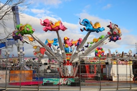A vibrant amusement ride featuring colorful cartoon elephants that are part of a rotating aerial ride structure. The elephants are painted in bright colors such as pink, green, blue, and red, with each one wearing a small hat. In the background, various carnival booths and decorations are visible, suggesting a lively fairground setting.