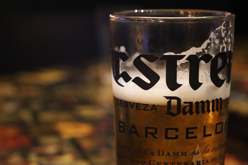 A close-up of a frothy glass of Estrella Damm beer with condensation on the outside, partially filled with clear golden liquid. The background appears to be dimly lit with blurred, colorful reflections.