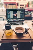 A cozy coffee shop setup with a laptop displaying photo editing software, placed on a table alongside a cup of tea and a small bottle of orange juice. The background shows a window view of a street with cars parked.